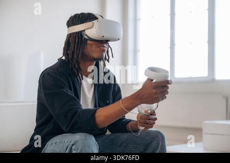 Relaxing by playing VR game, sitting. Black man in virtual reality glasses at home. Stock Photo