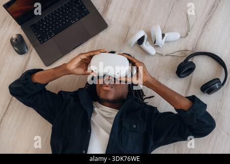 Computer nearby. Top view of man that is lying down on floor in virtual reality headset. Stock Photo