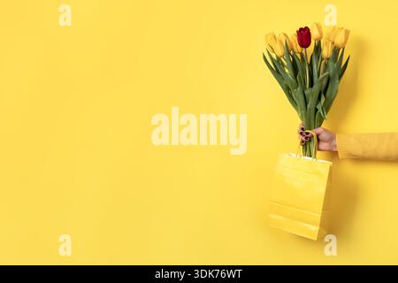 Woman’s hand holding yellow shopping bag with bouquet of yellow and red tulips against bright yellow background. Minimalist, copy space. Stock Photo