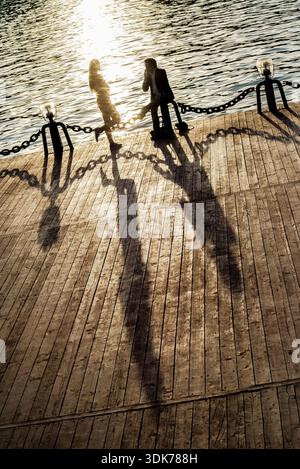 Two people standing by a chain barrier near water, casting long shadows on a paved surface. Stock Photo