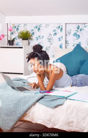 A young woman is lying on her bed, intently studying on her laptop. Surrounded by soft blankets and colorful pillows, she enjoys a lively atmosphere f Stock Photo