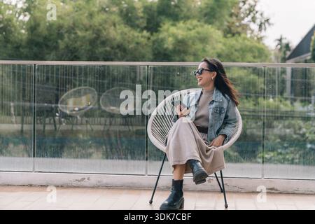 Young woman smiling while sitting on balcony using phone Stock Photo