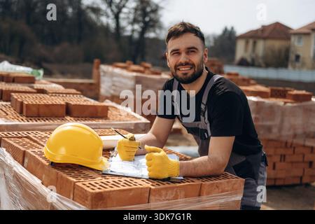 Positive facial expression, standing. Construction worker is at job with bricks outdoors. Stock Photo