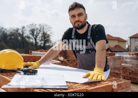 Positive facial expression, standing. Construction worker is at job with bricks outdoors. Stock Photo