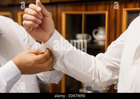 A man in a crisp white dress shirt carefully fastens his cufflinks, showcasing formal elegance and meticulous grooming against a warm wooden interior Stock Photo