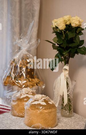 A beautifully arranged table features white roses in a glass vase alongside traditional round bread loaves wrapped in plastic, evoking a festive Easte Stock Photo