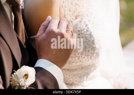 A tender close-up of a bride and groom holding hands, highlighting their wedding rings, elegant attire, and romantic connection during a sunny outdoor Stock Photo