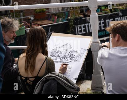 Group of artists sitting down sketching and drawing the view of a barge, church and buildings from the canal bridge in the Dutch city of Delft. Stock Photo