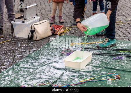 Street artist pours green bubble solution into container on pavement, tools and ropes around, showing preparation before performance. Urban scene Stock Photo