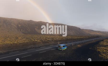 Aerial view of a vibrant blue van driving under a rainbow against the stark Icelandic landscape, the rugged mountains providing a dramatic backdrop, S Stock Photo