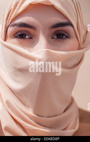 Female with dark hair, wearing a light hijab, gazes at the viewer with expressive eyes, creating a calm atmosphere against a soft, neutral backdrop || Model released Stock Photo