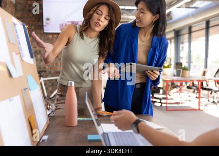 Diverse female coworkers examining pinned notes on cork board with tablet at coworking table. Collaboration, teamwork, innovation, creative space, modern office, productivity, professional || Model released Stock Photo