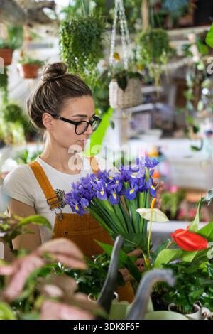 Surprised florist woman in glasses, wear orange overalls surrounded by houseplants, charmed with pleasant gift, looking at a bouquet of irises flowers. Indoor garden || Model released Stock Photo
