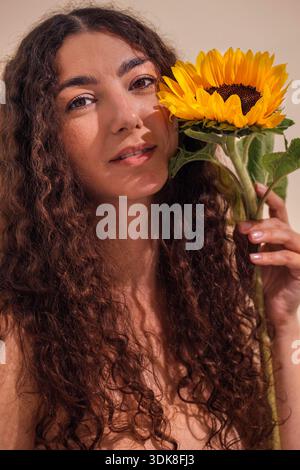 Female with curly hair gently holds sunflower, highlighting her natural beauty and connection to nature in a serene, warm environment filled with light || Model released Stock Photo