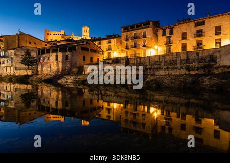 Nighttime River Town With Warm Lights And Arched Stone Bridge Reflecting In Calm Water, Valderrobres, Spain Stock Photo