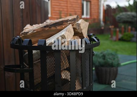 Firewood stacked in metal basket standing on outdoor patio near house. Wood logs prepared for heating, rustic fuel storage, natural material, backyard Stock Photo