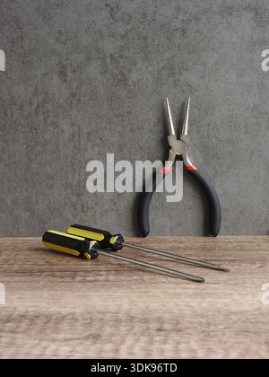 Different tools on a wooden table and a gray background, a round-nose tweezers with two screwdrivers with black and yellow handles very useful Stock Photo
