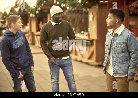 Three friends chatting and laughing in a festive outdoor christmas market setting, dressed casually, spending sparetime together, meeup, meeting each Stock Photo