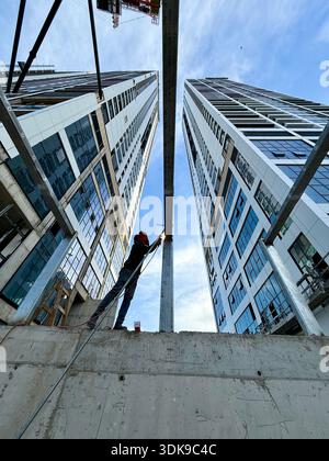 Construction worker on a ladder welding steel beams at a high-rise building site. Low-angle view of industrial labor against a modern skyscraper. Stock Photo