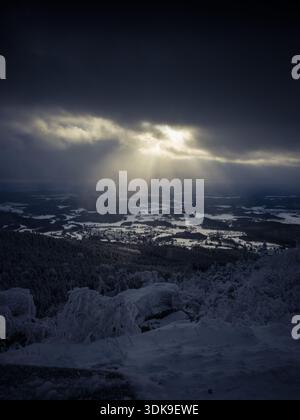 Sunbeams breaking through dark winter clouds above a snow-covered landscape viewed from Ještěd Mountain, creating a dramatic natural light display. Stock Photo