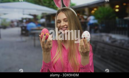 Young woman wearing bunny ears smiles outdoors while holding colorful easter eggs, embodying festive joy in a lively public setting. Stock Photo