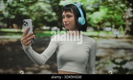 Young hispanic woman with cat ear headphones holds smartphone and beckons hand in forest; friendly invitation. Stock Photo