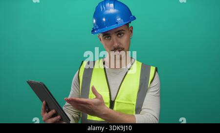 Young hispanic man in reflective vest and hardhat using tablet against isolated green background wall. Stock Photo