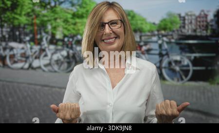 Smiling middle age woman beckons with hands on street in urban canal district; friendly invitation. Stock Photo