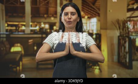 Woman wearing apron presses hands together in prayerlike gesture inside a warmly lit building with visible tables and lamps; calm welcome. Stock Photo