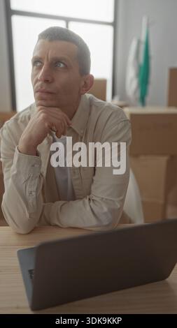 Man pondering in new apartment surrounded by moving boxes and laptop on wooden table with natural light filtering through large windows. Stock Photo
