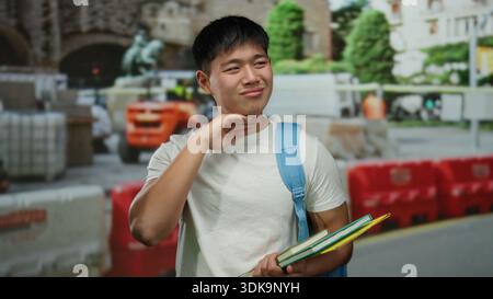 Young man holding books and feeling discomfort at construction site background with barriers and buildings in the distance. Stock Photo