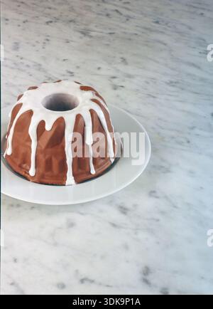A whole, round chocolate cake covered in smooth white icing , elegantly presented on a white plate against a white marble background. Stock Photo