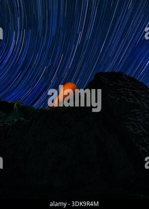 Orange plushie on a rocky summit beneath sweeping circular star trails in a deep blue night sky. Stock Photo