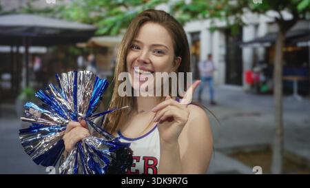 Young cheerleader woman holding blue metallic pompom points finger and smiles widely on street; enthusiasm. Stock Photo