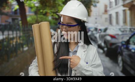 Chinese woman architect wearing hardhat holds blueprints and gives thumbs up on a vibrant street, symbolizing professionalism and confidence in an urb Stock Photo