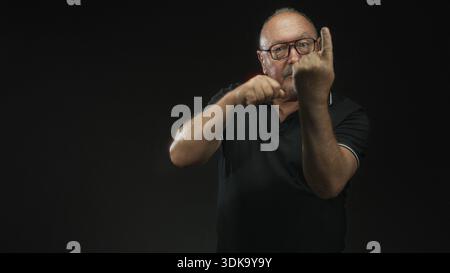 Man with clenched fists punching air in studio, wearing glasses and a black polo, showing knuckles and forearms against a dark backdrop; determination Stock Photo