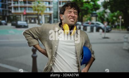 Hispanic man with headphones and hardhat stands on street clutching his back, showing discomfort while wearing casual clothing in an urban outdoor set Stock Photo