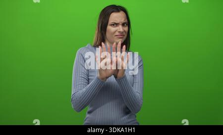 Woman with hands raised showing palms and grimace, recoil gesture, wearing blue sweater in studio against vivid green backdrop; disgust. Stock Photo