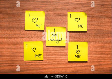 Five yellow sticky notes arranged on a wooden surface, featuring handwritten affirmations in black ink. Four notes display the message "I Love ME" wit Stock Photo