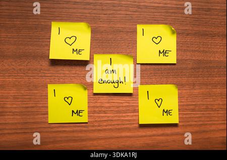Five yellow sticky notes arranged on a wooden surface, featuring handwritten affirmations in black ink. Four notes display the message "I Love ME" wit Stock Photo