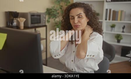Young hispanic woman in business attire sits at a computer desk in a modern office holding her neck with both hands; work fatigue. Stock Photo