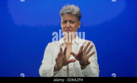 Woman holds palms forward in blue studio setting showing refusal posture with clear hands up stop gesture; disgust. Stock Photo