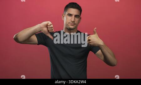 Young hispanic man giving thumbs up and thumbs down in a red studio wall setting; indecision doubt. Stock Photo