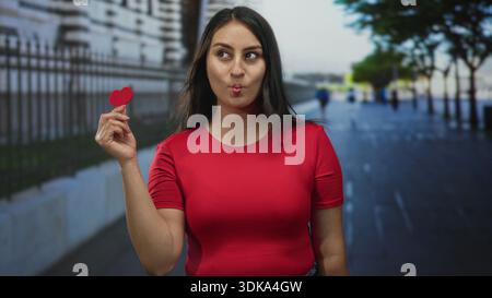 Young hispanic woman holds a small red paper heart between fingers and puckers lips into a fish face facing camera on street; playfulness. Stock Photo