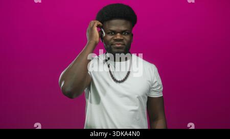 Man touching his ear with hand while wearing white t shirt and wooden bead necklace against magenta wall in studio; doubt. Stock Photo