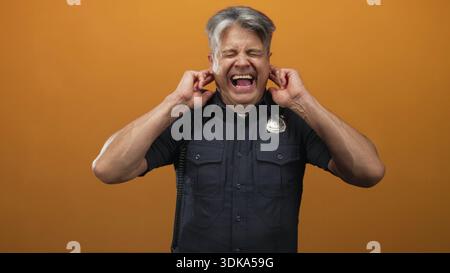 Police officer man plugs ears with fingers, grimacing and wincing in an orange studio with visible badge and radio cord; discomfort annoyance. Stock Photo