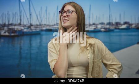 Blonde woman wearing glasses pinches nose beside boats on sunlit street pier over blue water; disgust. Stock Photo