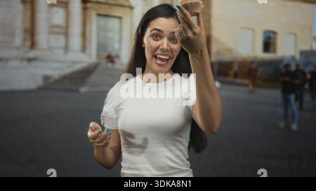 Young hispanic woman beckons with hand while holding muffin on street near historic building, smiling to camera; joy friendly invite. Stock Photo