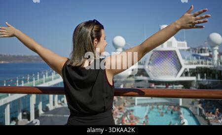 Woman standing with outstretched arms and hands on railing on cruise deck overlooking pool and sea; serenity travel escape. Stock Photo