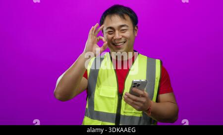 Man wearing safety vest using smartphone against vibrant pink background looks focused while texting an important message, embodying modern communicat Stock Photo